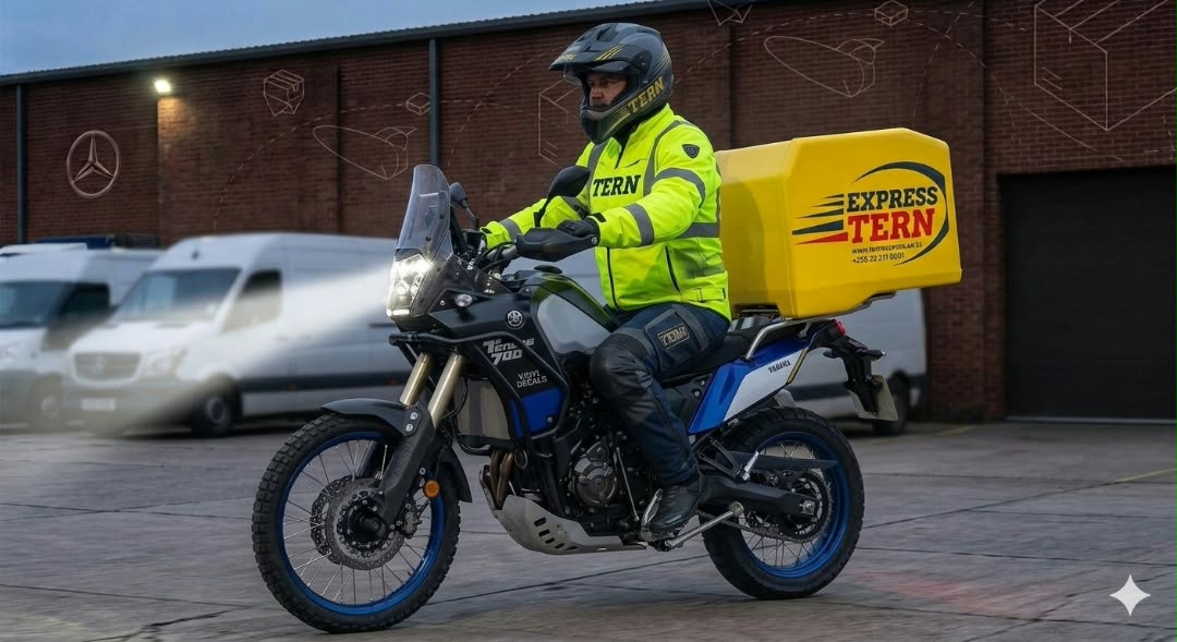 A Tern Express rider in a hi-vis jacket on a motorbike with a yellow delivery box, outside a depot with delivery vans